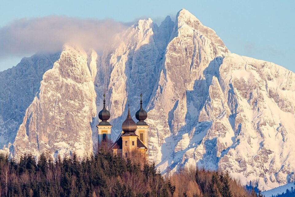 Eine malerische Berglandschaft mit schneebedeckten Gipfeln. Im Vordergrund steht eine schöne Kirche mit auffälligen Zwiebeltürmen.