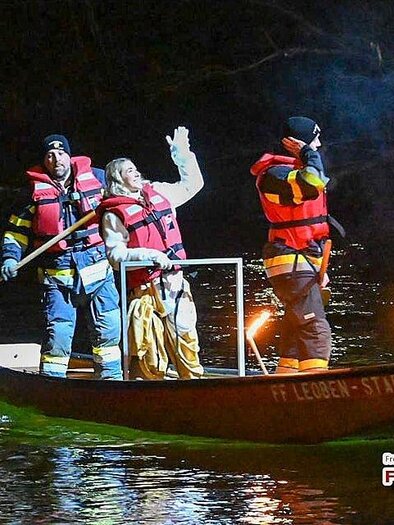 Three people in life jackets are riding in a boat on the water. They are holding torches in their hands and are out at night. | ©  FF Leoben-Stadt
