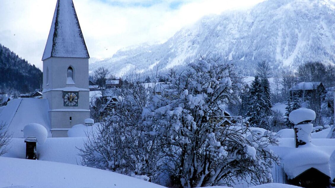 Eine verschneite Landschaft mit einem hohen Kirchturm. Im Hintergrund sind Berge und ein winterlicher Himmel zu sehen. | © TVB Ausseerland Salzkammergut_Pirker