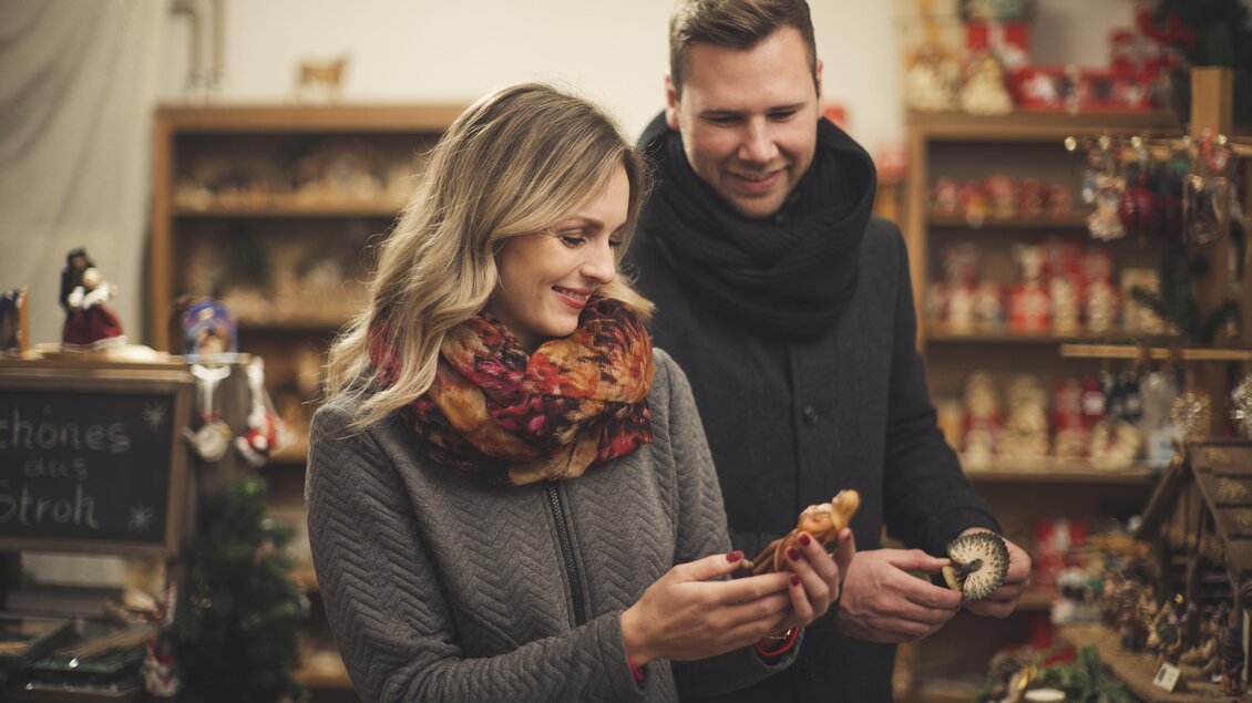 Ein Paar schaut sich liebevoll handgefertigte Figuren in einem festlich dekorierten Laden an. Die warme Atmosphäre und die Weihnachtsdekoration schaffen ein gemütliches Ambiente. | © Stadtgemeinde Feldbach I Bernhard Bergmann