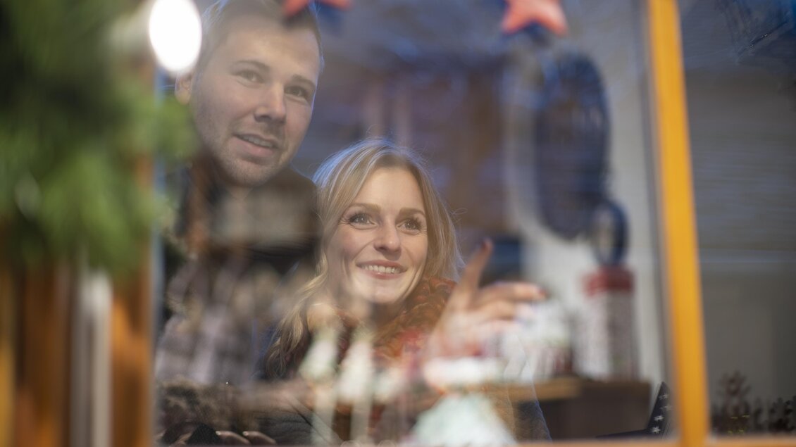 Ein Paar schaut durch ein Fenster auf festliche Dekorationen. Sie wirken glücklich und genießen die weihnachtliche Atmosphäre. | © Stadtgemeinde Feldbach I Bernhard Bergmann