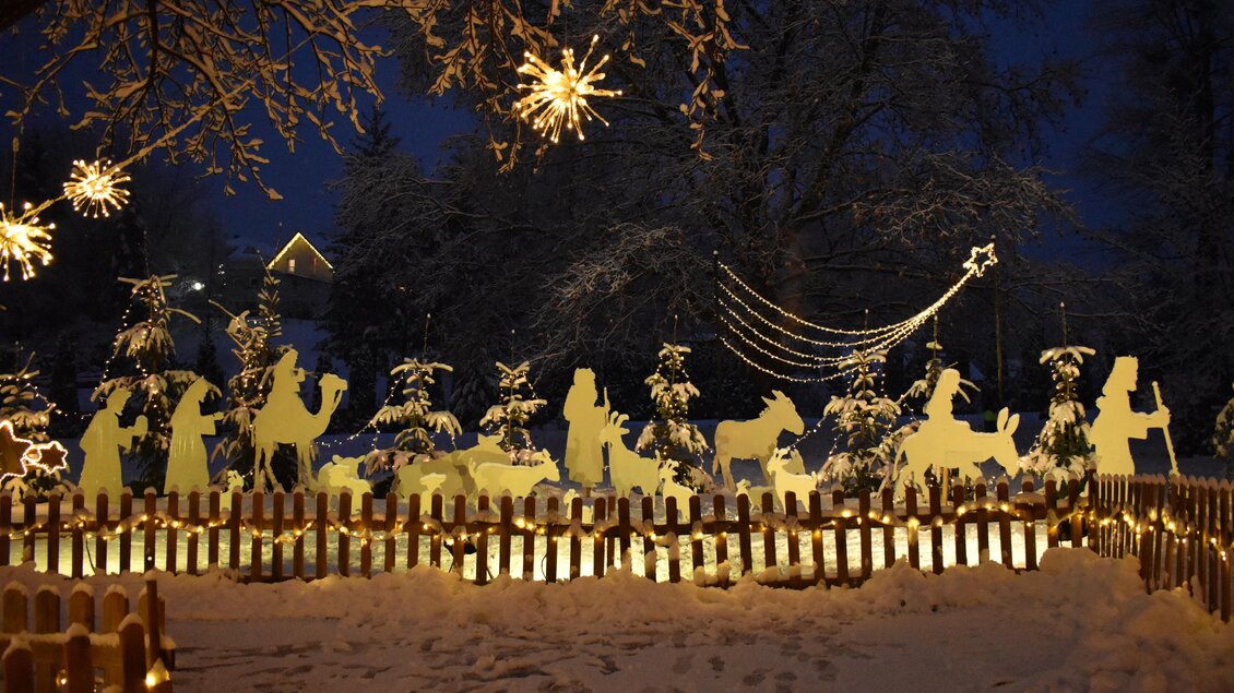 Eine winterliche Szenerie mit einer festlichen Weihnachtskrippe aus beleuchteten Figuren. Der Schnee reflektiert das Licht, während Schneeflocken und Sterne den Nachthimmel schmücken. | © Fam. Zwanzleitner