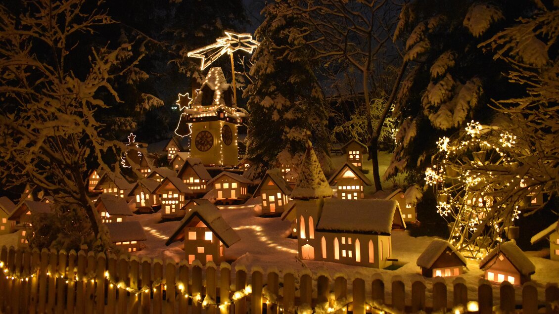 Eine festliche Winterlandschaft mit kleinen, beleuchteten Häusern im Schnee. Im Hintergrund steht eine Uhr mit einem hellen Stern und funkelnden Lichtern. | © Fam. Zwanzleitner