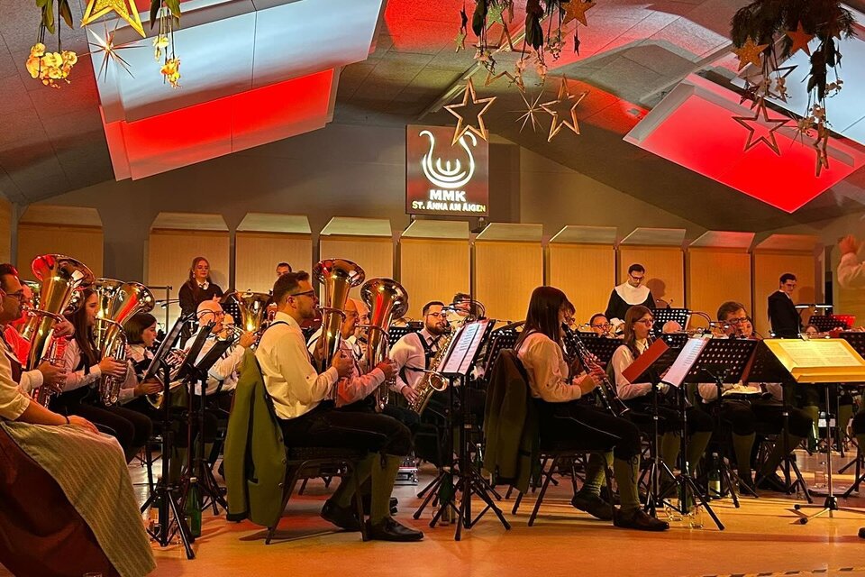 Ein Blasorchester spielt auf der Bühne. Der Raum ist festlich mit Dekorationen und buntem Licht gestaltet. | © Marktmusikkapelle St. Anna/A.