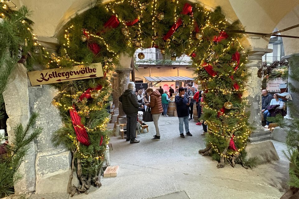 Eine festlich geschmückte Eingangspforte mit Lichtern und Tannenzweigen. Menschen stehen zusammen und genießen die Atmosphäre eines Weihnachtsmarktes. | © Thermen- & Vulkanland