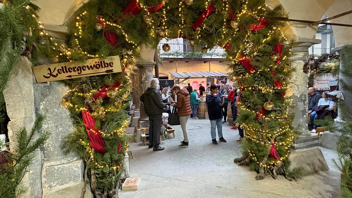 Eine festlich geschmückte Eingangspforte mit Lichtern und Tannenzweigen. Menschen stehen zusammen und genießen die Atmosphäre eines Weihnachtsmarktes. | © Thermen- & Vulkanland