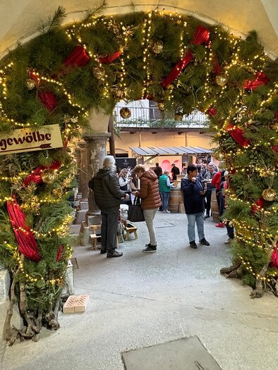A festively decorated entrance gate with lights and fir branches. People are gathered together and enjoying the atmosphere of a Christmas market. | © Thermen- & Vulkanland
