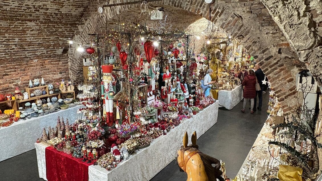 Ein festlich dekorierter Raum mit Weihnachtsständen und buntem Weihnachtsschmuck. Menschen betrachten die Ausstellung zwischen den Ständen. | © Thermen- & Vulkanland