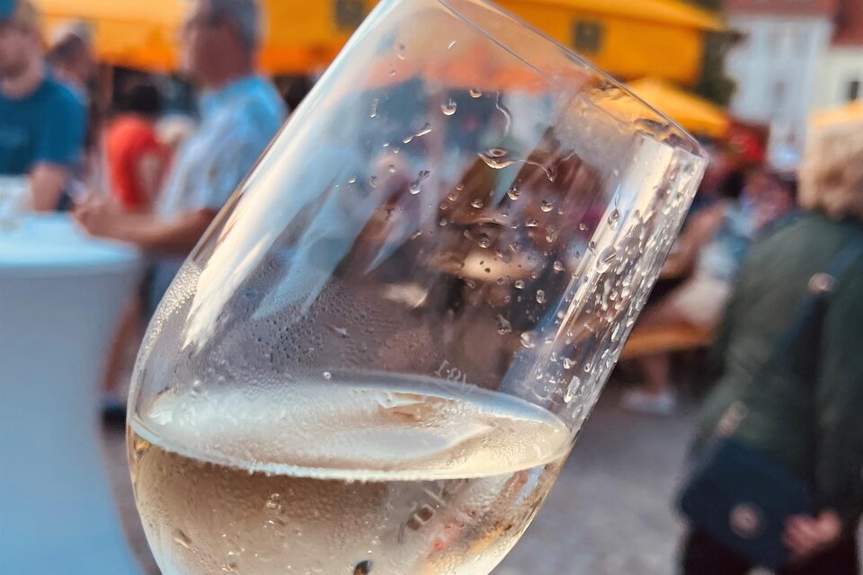 Ein Hand hält ein Glas mit sprudelndem Wein. Im Hintergrund sind Menschen und gelbe Markisen zu sehen. | © Stadtmarketing Fürstenfeld