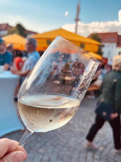 A hand holds a glass of sparkling wine. In the background, people and yellow awnings can be seen. | © Stadtmarketing Fürstenfeld