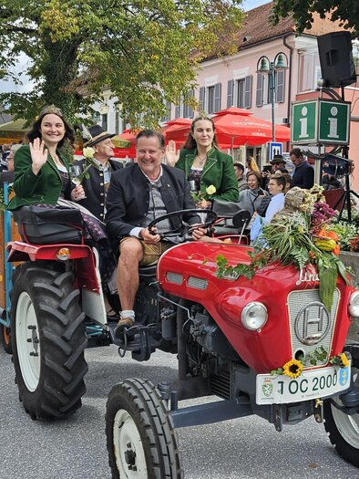 A festive parade featuring a red tractor with people dressed in traditional attire sitting on it. The floats are decorated with flowers and attract many spectators. | © Gemeinde Eibiswald