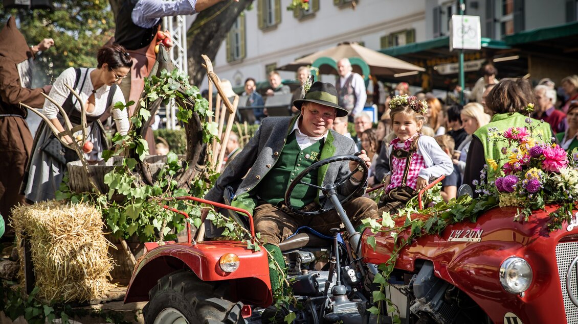 Eine festliche Parade mit einem Mann in traditioneller Tracht auf einem roten Traktor. Um ihn herum stehen fröhliche Menschen und dekorative Pflanzen. | © Achromatic Photography