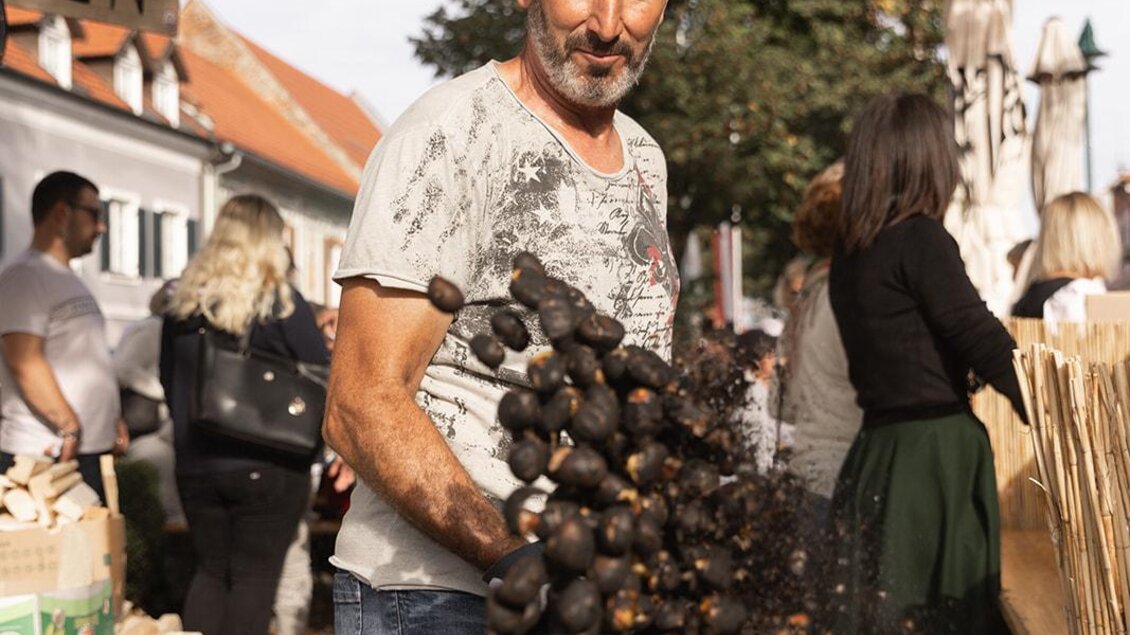 Ein Mann steht auf einem Markt und wirft frisch geröstete Kastanien in eine Pfanne. Im Hintergrund sind weitere Marktbesucher und Stände zu sehen. | © Achromatic Photography