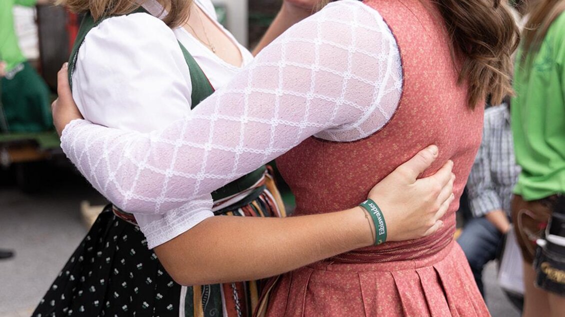 Zwei Frauen in traditioneller Tracht umarmen sich und lachen. Sie tragen Haarschmuck und stehen auf einer belebten Straße. | © Achromatic Photography
