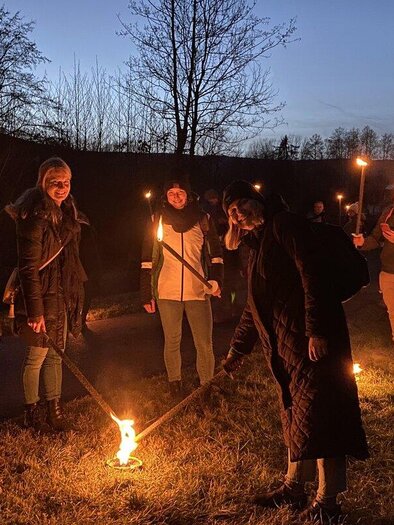 A group of people stands in the dark, holding torches in their hands. They are surrounded by trees, and the sky is dark blue. | © cdm - Claudia Dunst
