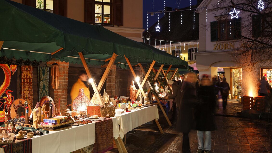 Ein festlicher Weihnachtsmarkt mit bunten Ständen und funkelnden Lichtern. Menschen schlendern in der Dämmerung und genießen die stimmungsvolle Atmosphäre.