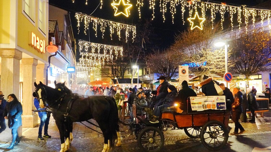 Ein festlicher Nachtmarkt mit vielen Menschen und einer Pferdekutsche. Über den Straßen hängen leuchtende Sterne und Lichter. | © Oststeiermark Tourismus