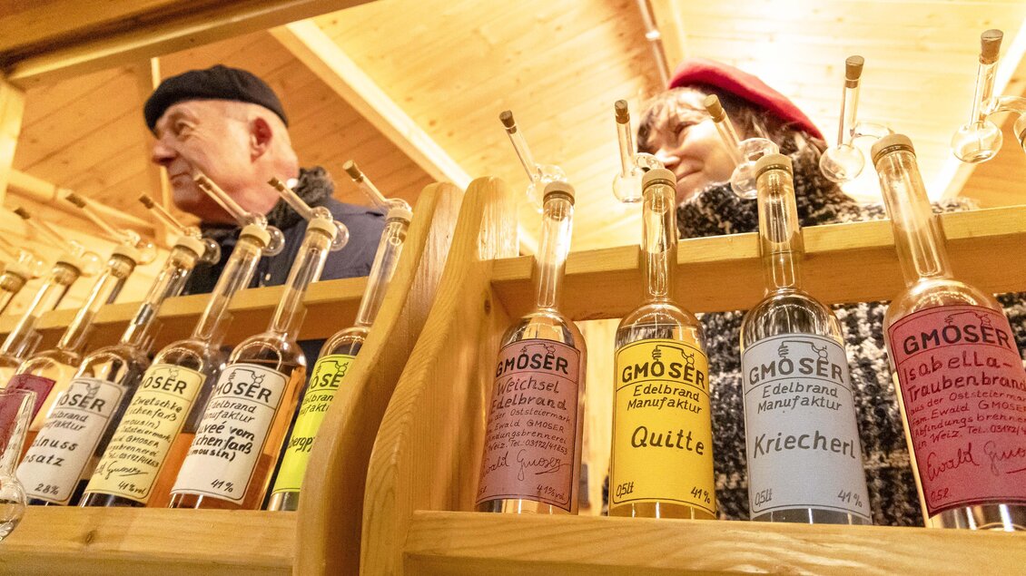 Zwei Personen stehen hinter einer Holztheke mit verschiedenen Flaschen. Die Flaschen enthalten Schnäpse mit bunten Etiketten.