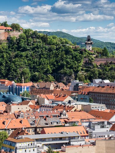 A city view with red roofs and green hills in the background. On the hill stands a historic castle, surrounded by blue sky and clouds. | © (c) Graz Tourismus - Harry Schiffer