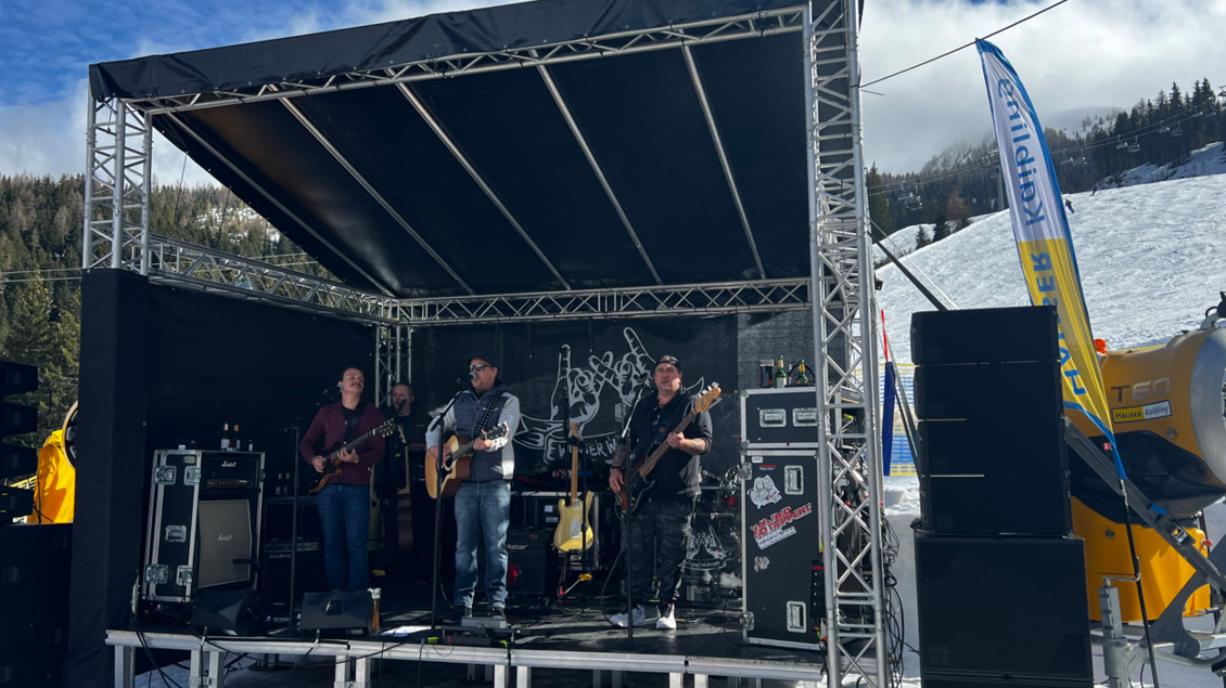 Eine Band spielt auf einer Bühne im Freien in einer schneebedeckten Landschaft. Im Hintergrund sind Bäume und ein blauer Himmel zu sehen. | © Hauser Kaibling
