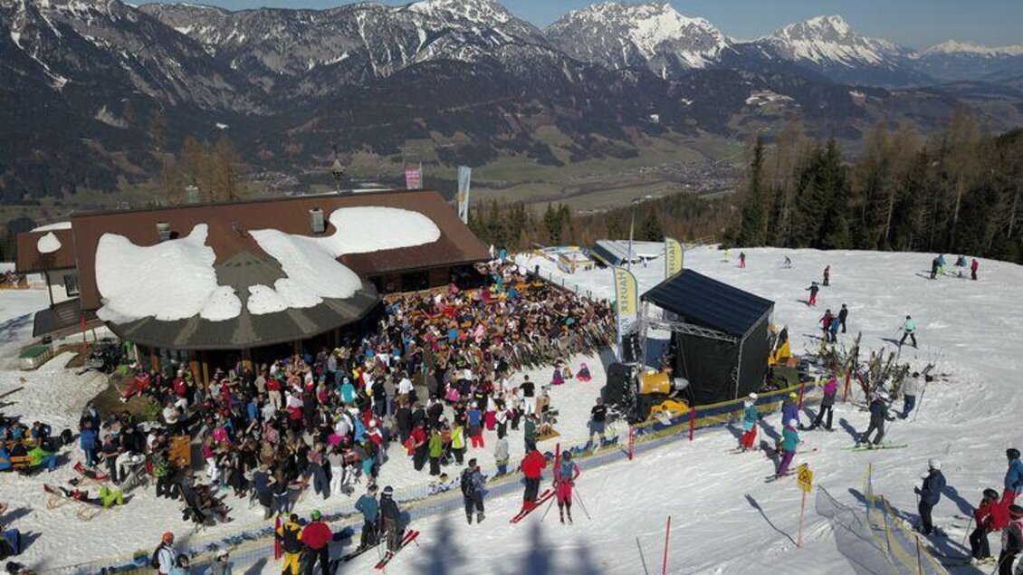 Eine lebhafte Skihütte mit vielen Menschen auf der Terrasse. Im Hintergrund erstrecken sich schneebedeckte Berge unter einem klaren Himmel.