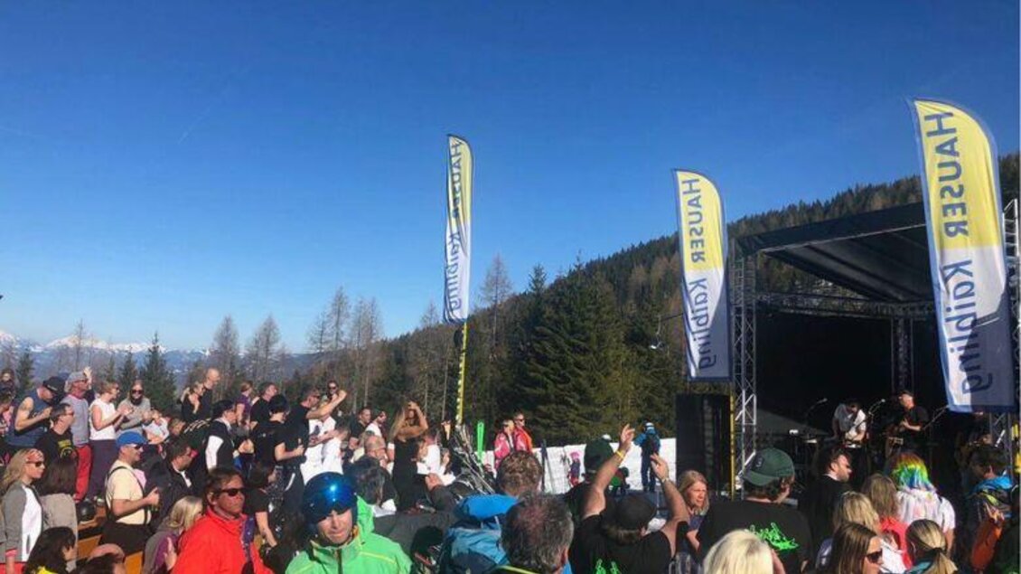 Ein Open-Air-Konzert in den Bergen mit vielen begeisterten Besuchern. Im Hintergrund sind Bäume und ein klarer Blauer Himmel zu sehen.