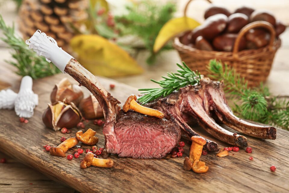 Ein köstliches Lammkarree, perfekt zubereitet, liegt auf einem Holzteller. Darum liegen frische Pfifferlinge und rosafarbene Pfefferkörner. | © Foto Fotolia