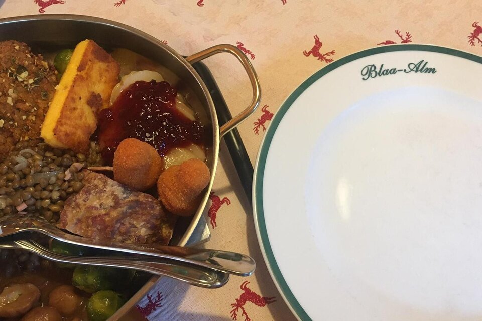Ein Teller mit verschiedenen Gerichten, darunter Fleisch, Gemüse und Beilagen. Daneben liegt ein leerer Teller mit der Aufschrift „Blaa-Alm“. | © Tanja Kirchschlager