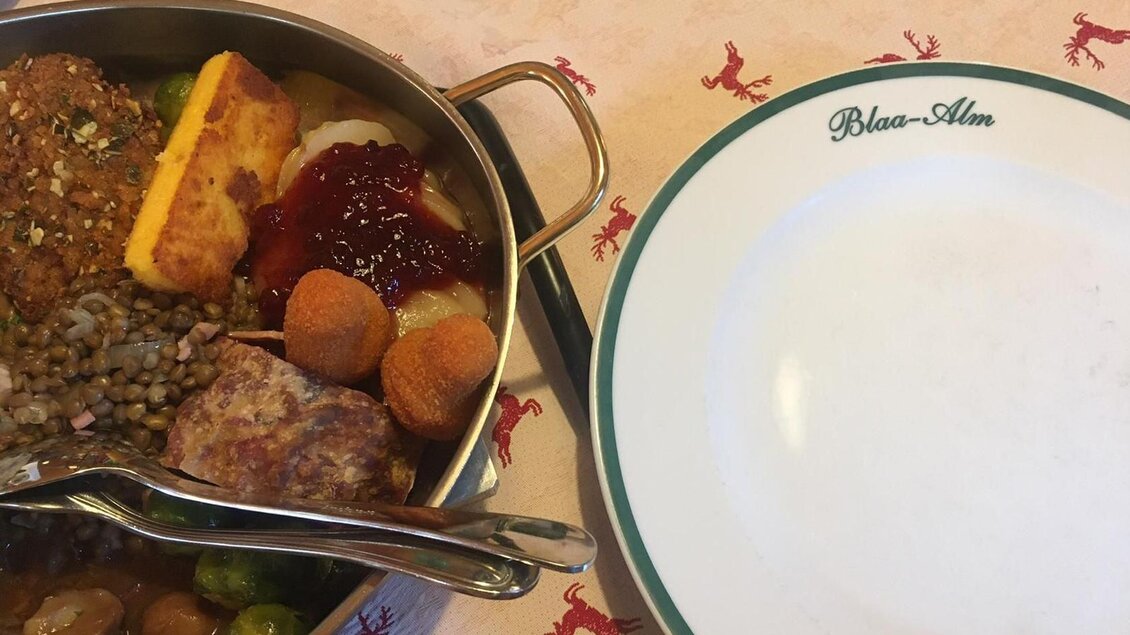 Ein Teller mit verschiedenen Gerichten, darunter Fleisch, Gemüse und Beilagen. Daneben liegt ein leerer Teller mit der Aufschrift „Blaa-Alm“. | © Tanja Kirchschlager