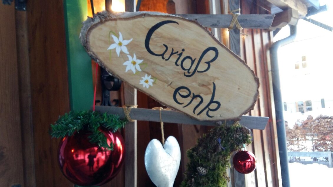 Ein handgefertigtes Holzschild mit der Aufschrift „Griaß enk“ und dekorativen Elementen wie Weihnachtsbaumkugeln und grünen Zweigen. Das Bild vermittelt eine gemütliche, festliche Atmosphäre. | © Petra Kirchschlager