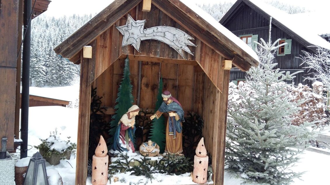 Eine Weihnachtskrippe steht in einem Holzstall, umgeben von schneebedeckten Bäumen. Maria und Josef betrachten das neugeborene Kind in der Mitte, während die Szene von winterlicher Schönheit umgeben ist. | © Petra Kirchschlager