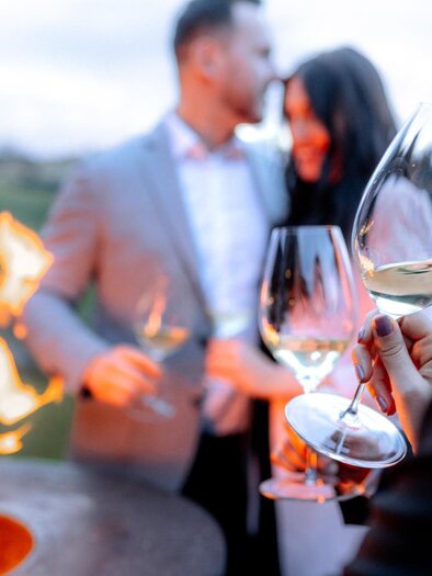 An elegant outdoor celebration with cheerful people holding wine glasses. In the background, a campfire can be seen, creating a cozy atmosphere. | © Daniel Obersberger