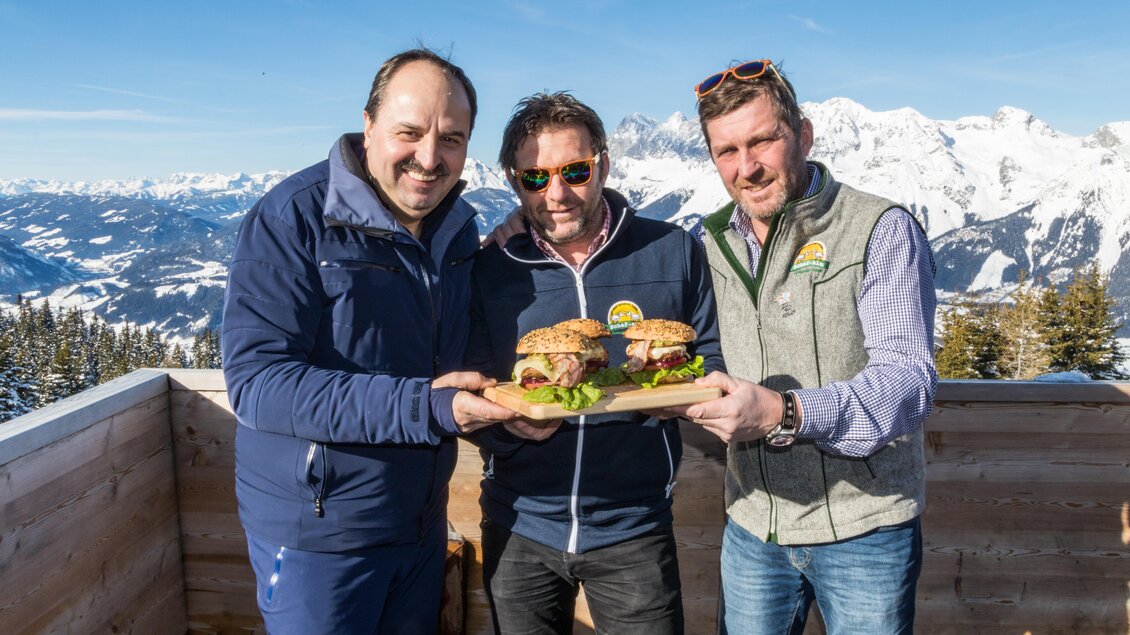 Drei Männer stehen in den Bergen und halten große Burger. Im Hintergrund sind schneebedeckte Berge und blauer Himmel zu sehen. | © Planai/Alexander Klünsner