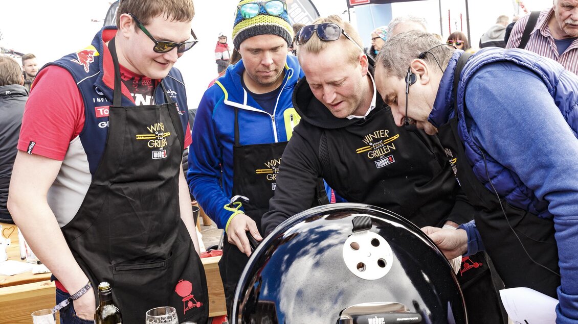 Eine Gruppe von Männern in Schürzen steht um einen Grill und diskutiert. Im Hintergrund sind Zuschauer zu sehen, die an einem Event teilnehmen. | © Christoph Buchegger