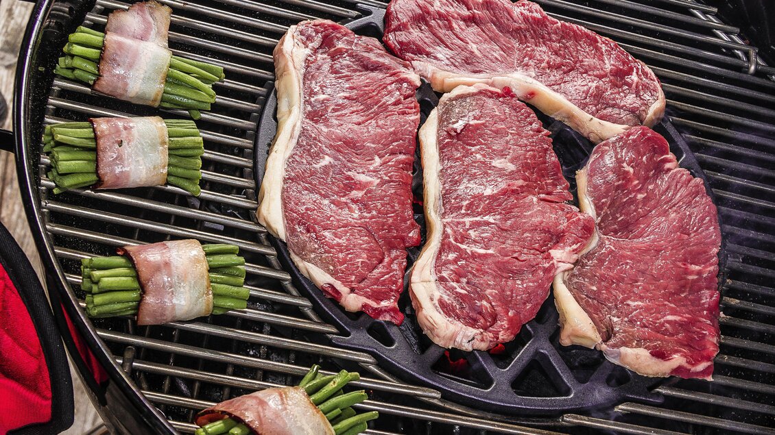 Saftige Steaks und mit Speck umwickelte grüne Bohnen liegen auf einem Grill. Die Szene vermittelt eine einladende Atmosphäre für ein Grillfest. | © Christoph Buchegger