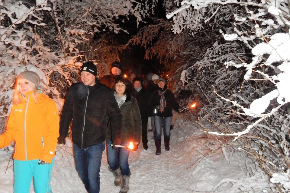 Eine Gruppe von Menschen wandert bei Nacht durch einen schneebedeckten Wald. Sie halten Fackeln in den Händen, die einen warmen Schein im kalten, weißen Umfeld erzeugen. | © Gemeinde Mitterberg-St. Martin