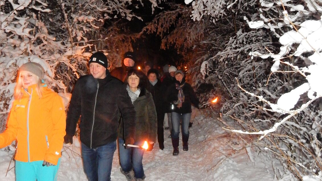 Eine Gruppe von Menschen wandert bei Nacht durch einen schneebedeckten Wald. Sie halten Fackeln in den Händen, die einen warmen Schein im kalten, weißen Umfeld erzeugen. | © Gemeinde Mitterberg-St. Martin