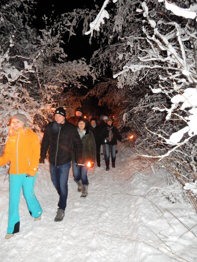 Eine Gruppe von Menschen wandert bei Nacht durch einen schneebedeckten Wald. Sie halten Fackeln in den Händen, die einen warmen Schein im kalten, weißen Umfeld erzeugen. | © Gemeinde Mitterberg-St. Martin