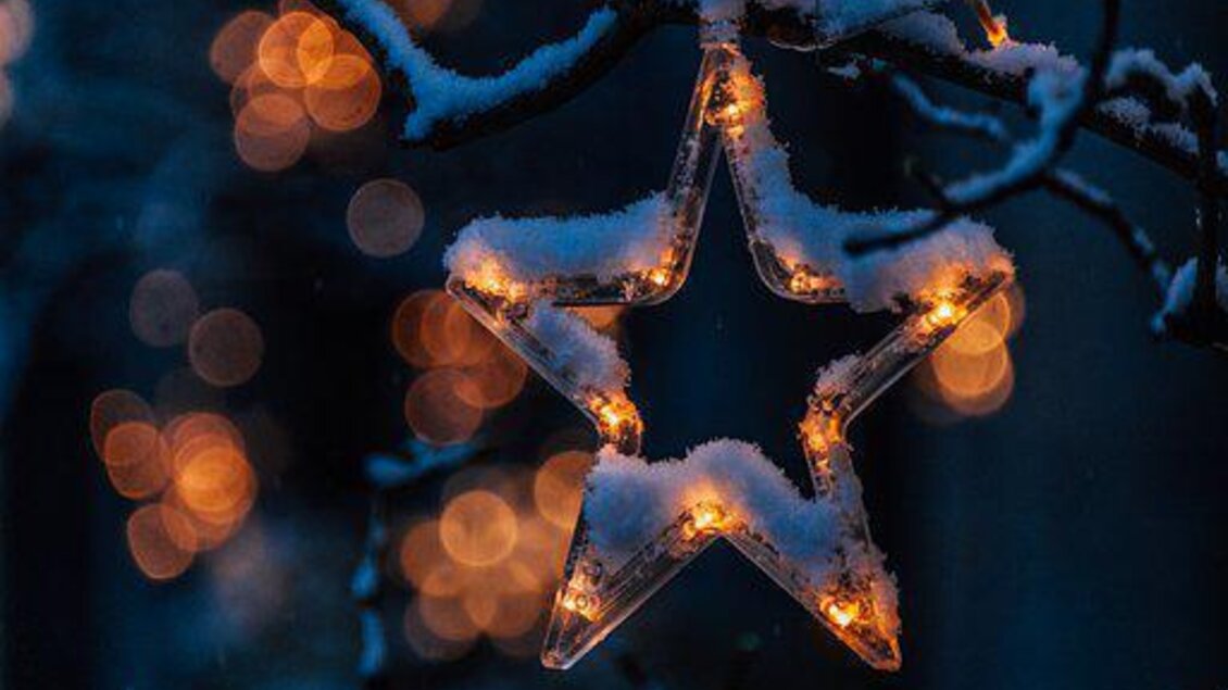 Ein leuchtender Weihnachtsstern, bedeckt mit Schnee, hängt an einem Ast. Im Hintergrund funkeln warmweiße Lichter. | © Pixabay 