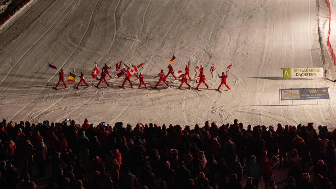 Eine Gruppe von Menschen in roten Anzügen marschiert auf einer verschneiten Piste. Im Hintergrund ist eine große Menschenmenge zu sehen. | © Michael Simonlehner