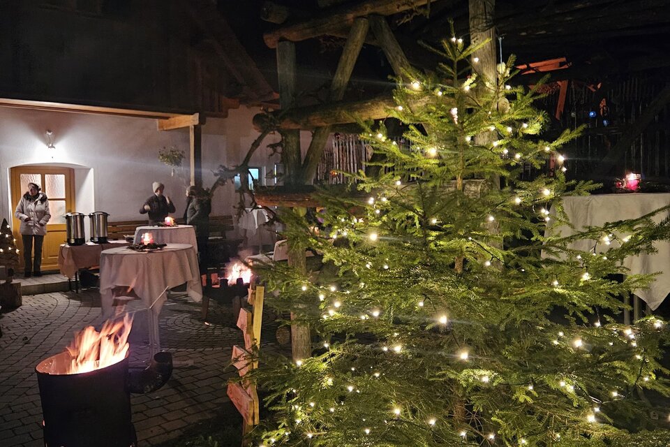 Ein festlich beleuchteter Weihnachtsbaum steht in einem gemütlichen Außenbereich. Im Hintergrund sind Menschen und ein Lagerfeuer zu sehen. | © Bäuerinnen Weinitzen