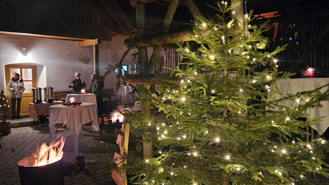Ein festlich beleuchteter Weihnachtsbaum steht in einem gemütlichen Außenbereich. Im Hintergrund sind Menschen und ein Lagerfeuer zu sehen. | © Bäuerinnen Weinitzen