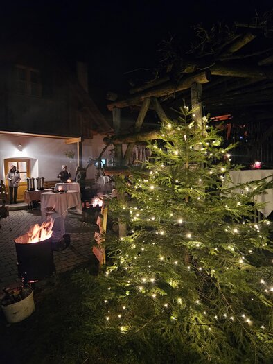 A festively lit Christmas tree stands in a cozy outdoor area. In the background, people and a campfire can be seen. | © Bäuerinnen Weinitzen