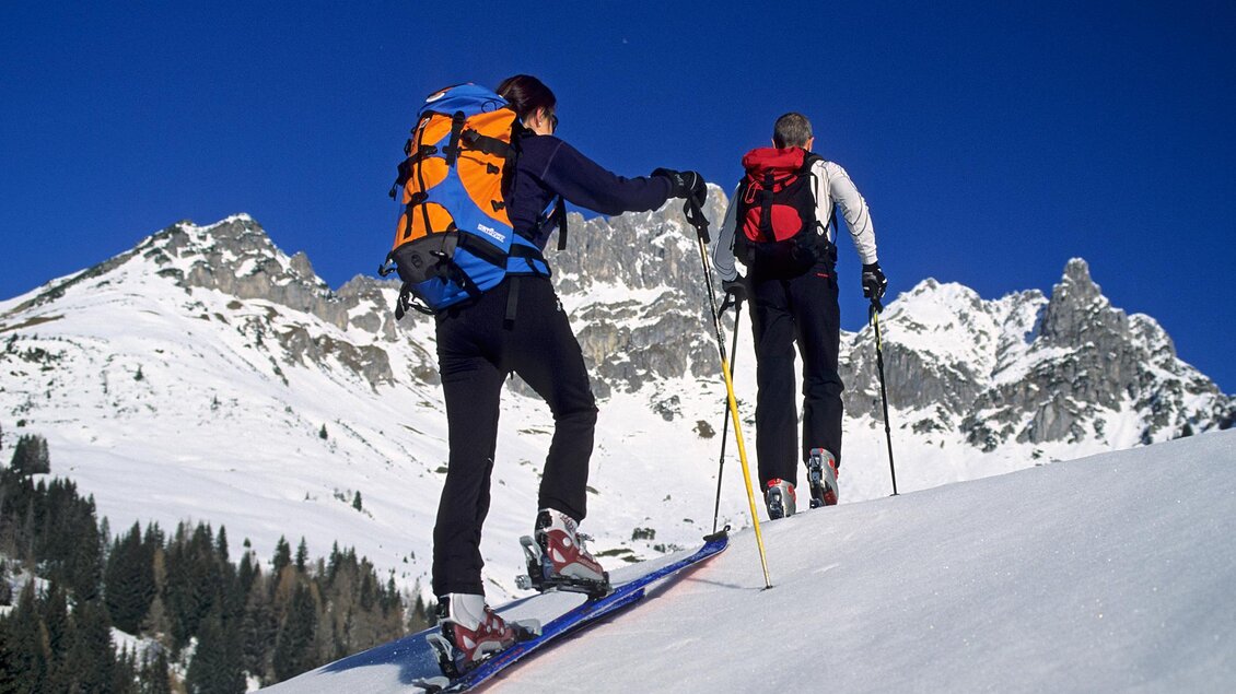 Zwei Personen ziehen mit Skiern einen schneebedeckten Hang hinauf. Im Hintergrund sind beeindruckende Berge und ein klarer blauer Himmel zu sehen. | © Alpincenter