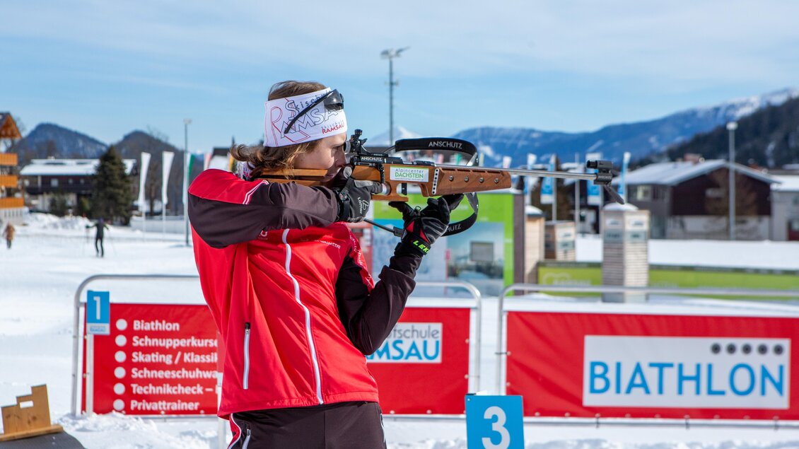 Eine Sportlerin im roten Outfit steht auf Schnee und zielt mit einem Gewehr auf ein Ziel. Im Hintergrund sind Skihütten und eine Biathlon-Arena zu sehen. | © Alpincenter