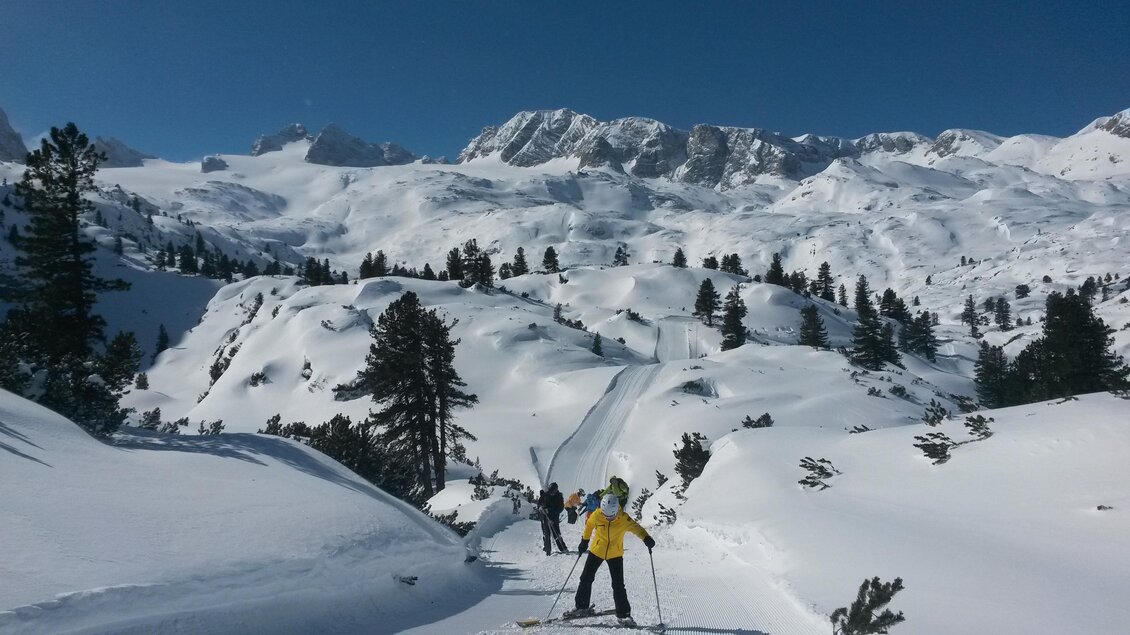 Eine schneebedeckte Landschaft mit Skifahrern auf einem schmalen Weg. Im Hintergrund erheben sich majestätische Berge unter einem strahlend blauen Himmel. | © Alpincenter