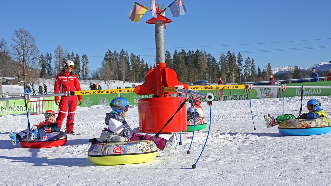 Eine Gruppe von Kindern sitzt in bunten Schläuchen auf einer verschneiten Piste. Im Hintergrund steht ein Skilehrer und es gibt eine klare blaue Himmel. | © Alpincenter