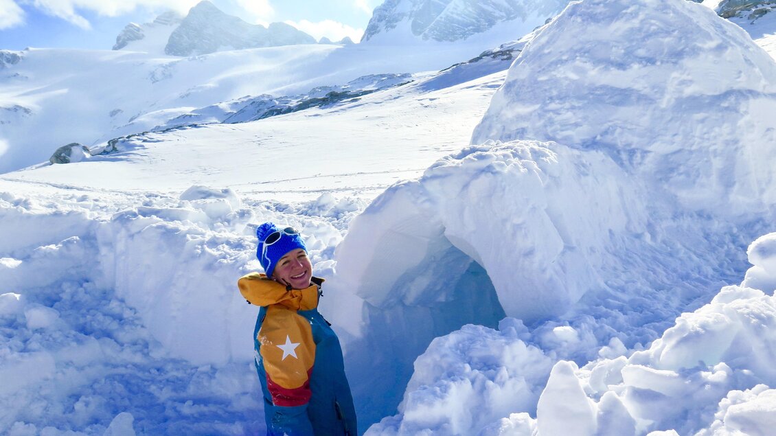 Eine Person steht vor einem Iglu in einer verschneiten Landschaft. Im Hintergrund sind Berge und ein klarer Himmel zu sehen. | © Heli Rettensteiner