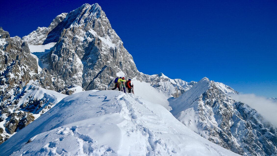 Eine Gruppe von Bergsteigern steht auf dem schneebedeckten Gipfel eines Berges. Im Hintergrund sind majestätische Berge und ein klarer, blauer Himmel zu sehen. | © Heli Rettensteiner