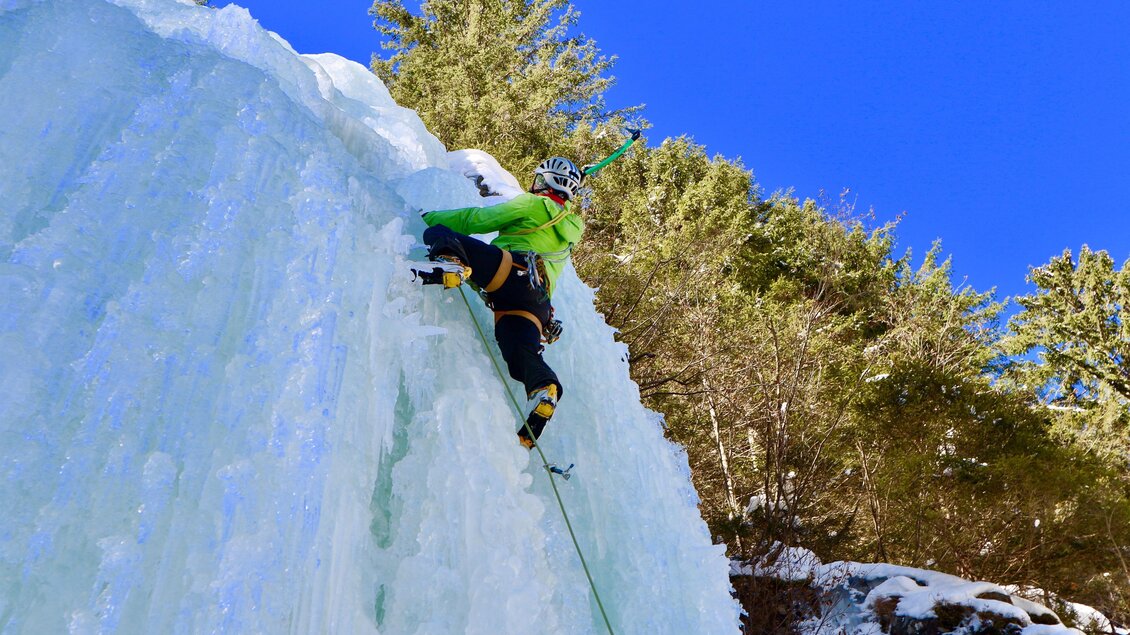 Ein Eiskletterer klettert eine gefrorene Wand mit einem Seil. Im Hintergrund sind grüne Bäume und ein klarer blauer Himmel sichtbar. | © Heli Rettensteiner